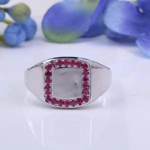 May include: A silver ring with a square center, outlined with small, round, red gemstones. The ring is set against a white background with blue and purple floral accents.