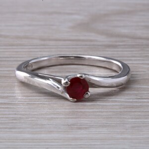 以下が含まれることがあります： 中央に丸い赤いルビーの宝石が1つセットされたシルバーリング。バンドはカーブしており、シンプルなデザインです。