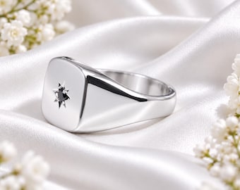 May include: A polished silver signet ring featuring a square face with rounded corners. A small, dark, round gemstone is set in the centre of the face, surrounded by an engraved starburst pattern with eight distinct points.