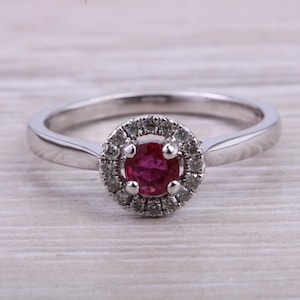 May include: A silver ring with a round, red gemstone center, surrounded by a halo of smaller, clear stones. The ring band is smooth and polished. The jewelry is set against a light wood grain background.