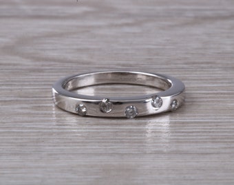 May include: A silver band ring with six small, clear gemstones evenly spaced around the top. The ring has a flat, polished surface and sits on a light-colored wooden surface. The ring is a simple, elegant design.