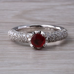 May include: A silver ring with a red gemstone surrounded by small diamonds. The band is covered in small diamonds.