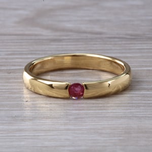 May include: A simple gold band ring with a single red gemstone set in the center.