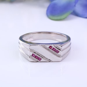 May include: A silver ring with a rectangular face, featuring three small, red gemstones. The ring has a textured design with diagonal lines and is set against a white background.