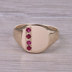 Könnte beinhalten: Ein goldener Siegelring mit einer glatten ovalen Oberseite und einer vertikalen Reihe von fünf roten Rubin-Steinen.