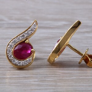 Op de afbeelding: Een paar gouden oorbellen met een robijn-edelsteen en een halo van diamanten. De oorbellen hebben een gebogen ontwerp en een oorbelsteker.