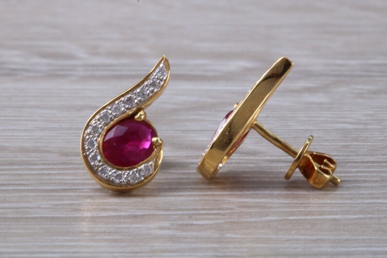 Op de afbeelding: Een paar gouden oorbellen met een robijn-edelsteen en een halo van diamanten. De oorbellen hebben een gebogen ontwerp en een oorbelsteker.