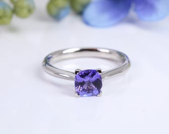 Puede incluir: Un anillo plateado con una gema cuadrada de color púrpura vibrante. El anillo está sobre una superficie blanca, con acentos florales verdes y azules borrosos en el fondo. La banda del anillo es lisa y pulida.