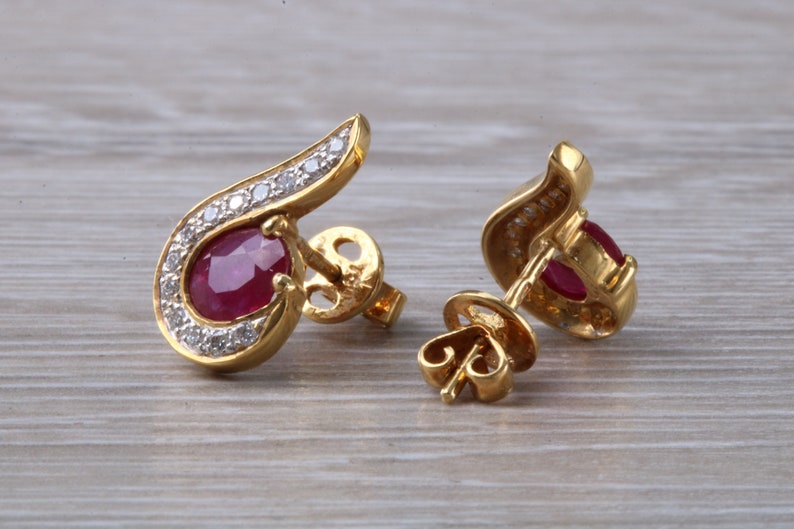 Op de afbeelding: Een paar gouden oorbellen met een rode robijn edelsteen en een halo van diamanten. De oorbellen hebben een gebogen ontwerp en een oorbelsteker.