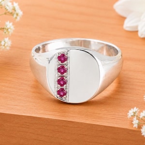 May include: A polished silver signet ring featuring a vertical channel set with four small, round, vibrant red rubies. The ring has a smooth, reflective surface on the right side of the face, contrasting with the gemstone-adorned left side.