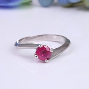 May include: A silver ring with a vibrant red gemstone. The ring features a sleek, modern design with a single, round-cut ruby set in a four-prong setting. The band curves gracefully, creating an elegant and eye-catching piece of jewelry.
