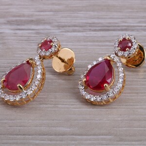 May include: A pair of gold earrings featuring a teardrop-shaped ruby gemstone surrounded by a halo of smaller diamonds.