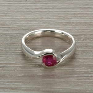 May include: A silver ring with a red ruby gemstone set in a curved band.