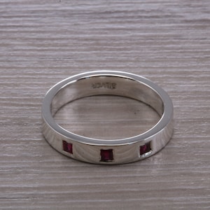 May include: A silver band ring with three small, square-shaped ruby stones set into the band.