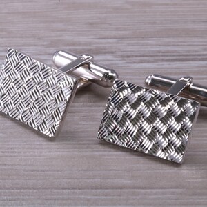 May include: A pair of silver cufflinks with a textured, woven pattern. The cufflinks are rectangular in shape and have a polished finish.