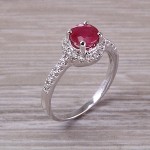May include: A silver ring with a red ruby gemstone surrounded by a halo of smaller diamonds. The band is also set with smaller diamonds.