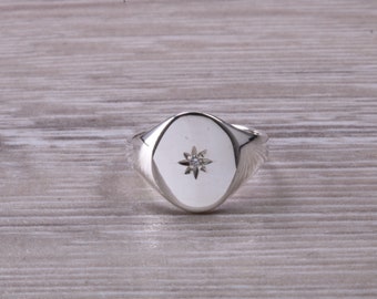 May include: A silver signet ring with a smooth oval top featuring a small, clear diamond set in a star shape.