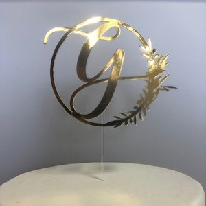 Peut inclure: Décor de gâteau en or avec une lettre G dans un cercle et un motif feuillu.