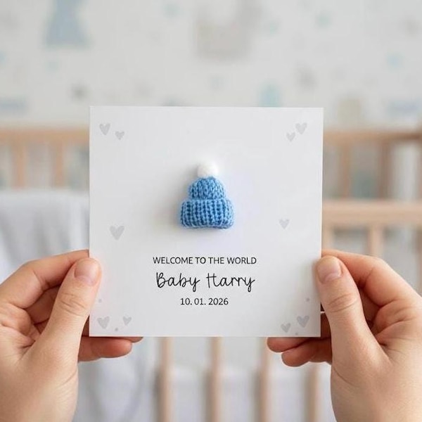 May include: A white card with a blue knitted baby hat and the text "WELCOME TO THE WORLD Baby Harry 10.01.2026". The card has small gray heart shapes. The hat has a white pom-pom.