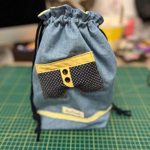 Może przedstawiać: Niebieska torba sznurkowa z denimu z żółtym wykończeniem i kieszenią w czarne i białe kropki. Kieszeń ma żółte wykończenie i dwa żółte guziki.