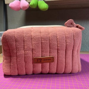 Może przedstawiać: Różowa kosmetyczka z pikowanego materiału z brązową skórzaną metką z napisem "ręcznie robione".