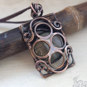 May include: A copper wire wrapped pendant with a green gemstone in the center. The pendant is rectangular in shape and has a delicate, intricate design. The pendant is hanging from a brown leather cord.