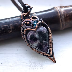 Pode incluir: Um pingente envolto em fio de cobre oxidado com uma pedra em forma de coração com padrões manchados em roxo, preto e branco, com um pequeno cristal azul. O pingente está pendurado num cordão escuro e inclui o texto Artarina.