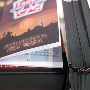 Puede incluir: Una baraja de tarot con cartas de bordes negros y un manual de instrucciones. La portada del manual muestra una ilustraci&oacute;n de un paisaje al atardecer, el texto "TAROT BY THE Neon Light" y "DESIGN and MANUAL by NICK RIBERA" con la p&aacute;gina 23.