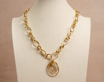 Puede incluir: Un collar de cadena en tono dorado con una serie de eslabones circulares y ovalados entrelazados. El collar incluye un colgante central compuesto por tres anillos ovalados texturizados anidados que cuelgan de un aro.