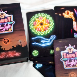 Puede incluir: Una baraja de tarot Tarot by the Neon Light de Chain Assembly con una caja negra que muestra un paisaje al atardecer y texto estilo ne&oacute;n. Varias cartas de tarot est&aacute;n desplegadas, mostrando ilustraciones de ne&oacute;n de s&iacute;mbolos.