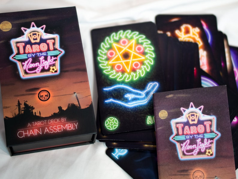 Puede incluir: Una baraja de tarot Tarot by the Neon Light de Chain Assembly con una caja negra que muestra un paisaje al atardecer y texto estilo ne&oacute;n. Varias cartas de tarot est&aacute;n desplegadas, mostrando ilustraciones de ne&oacute;n de s&iacute;mbolos.
