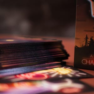 Puede incluir: Una baraja de cartas de tarot con bordes oscuros apilada junto a una caja que muestra la silueta de un paisaje bajo un sol rojo brillante. La caja muestra el texto Neon, A TAROT DECK y CHAIN AS. Las cartas presentan ilustraciones estilo ne&oacute;n.