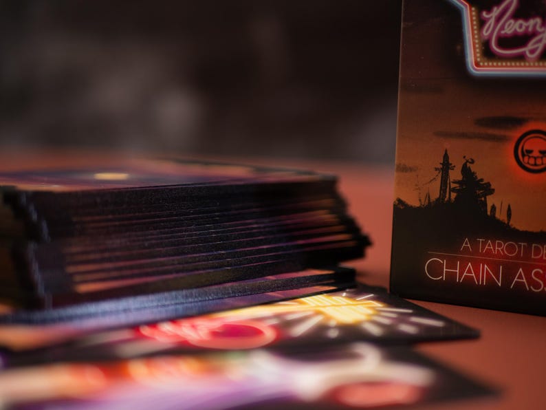 Puede incluir: Una baraja de cartas de tarot con bordes oscuros apilada junto a una caja que muestra la silueta de un paisaje bajo un sol rojo brillante. La caja muestra el texto Neon, A TAROT DECK y CHAIN AS. Las cartas presentan ilustraciones estilo ne&oacute;n.