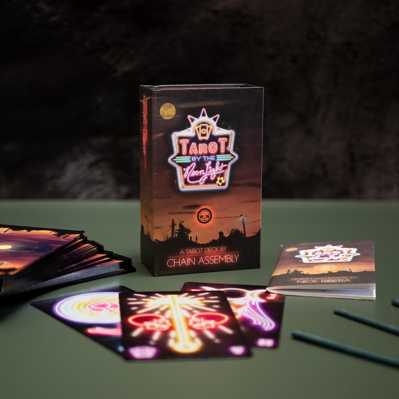 Puede incluir: Una caja de baraja Tarot by the Neon Light con un gr&aacute;fico de letrero de ne&oacute;n brillante, un paisaje al atardecer y el texto "A Tarot Deck by Chain Assembly". Varias cartas de tarot individuales con ilustraciones de ne&oacute;n est&aacute;n extendidas.