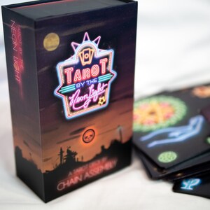 Puede incluir: Una caja rectangular de cartas de tarot con un fondo de atardecer oscuro y un letrero estilo ne&oacute;n que dice Tarot by the Neon Light. La caja presenta una silueta de ciudad y el texto Chain Assembly. Varias cartas de tarot negras est&aacute;n desplegadas.