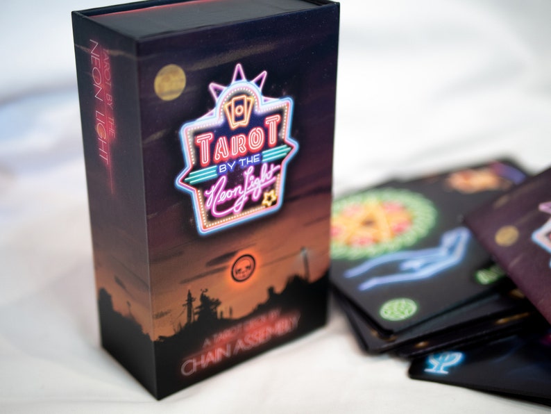 Puede incluir: Una caja rectangular de cartas de tarot con un fondo de atardecer oscuro y un letrero estilo ne&oacute;n que dice Tarot by the Neon Light. La caja presenta una silueta de ciudad y el texto Chain Assembly. Varias cartas de tarot negras est&aacute;n desplegadas.