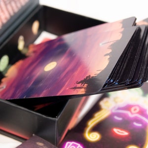 Puede incluir: Una caja de almacenamiento negra que contiene un mazo de cartas rectangulares. La carta superior presenta un paisaje de atardecer con una luna amarilla, mientras que otra carta muestra una ilustraci&oacute;n estilo ne&oacute;n de un rostro con contornos brillantes.