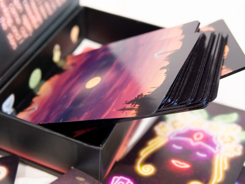 Puede incluir: Una caja de almacenamiento negra que contiene un mazo de cartas rectangulares. La carta superior presenta un paisaje de atardecer con una luna amarilla, mientras que otra carta muestra una ilustraci&oacute;n estilo ne&oacute;n de un rostro con contornos brillantes.