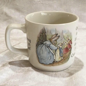 May include: A cream-colored ceramic mug featuring a printed illustration of a rabbit in a blue coat carrying a bundle, standing next to a smaller rabbit. The mug includes printed text segments from a storybook on both sides of the illustration.