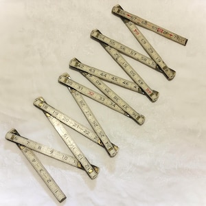 May include: A vintage folding wooden ruler with a white finish and black numerical markings. The ruler is partially unfolded, displaying segments with numbers ranging from 1 to 71 inches. The joints are metallic, showing signs of wear and age.