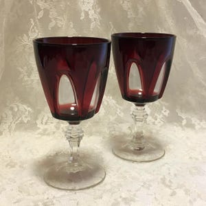 May include: Two matching wine glasses featuring deep red bowls with clear glass arched cutouts. Each glass has a clear, multi-faceted stem and a circular base. The design showcases a contrast between the dark crimson glass and the transparent sections.