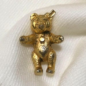 May include: A gold-toned metal bear pendant featuring a textured surface, rounded ears, and a small circular detail on the chest. The bear is depicted in a standing position with arms extended outward and a small loop at the top for attachment.