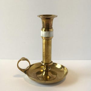 May include: A brass chamberstick candle holder featuring a circular base with a side handle and a central stem. The stem is accented with a band of white mother-of-pearl inlay. The metal surface has a polished finish with visible golden tones.