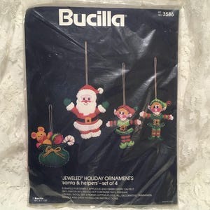May include: A Bucilla kit number 3586 for four jeweled holiday ornaments featuring Santa and his helpers. The package displays a felt Santa, two elves, and a gift bag with a mouse, all with hanging cords, against a dark blue background.
