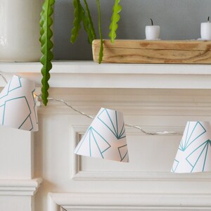 May include: A string of white paper cone lights with a teal geometric pattern. The lights are strung across a white mantel.
