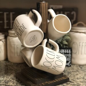 May include: A wooden mug rack with four white ceramic mugs hanging from it. The mugs have black lettering that reads "Self Made", "Soccer Mom", and "Rae Dunn".