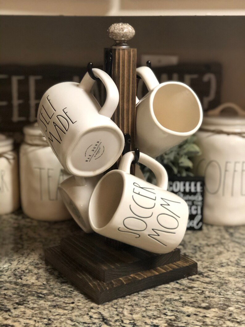 May include: A wooden mug rack with four white ceramic mugs hanging from it. The mugs have black lettering that reads "Self Made", "Soccer Mom", and "Rae Dunn".