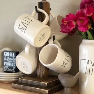 May include: A wooden mug rack with four white ceramic mugs hanging from it. The mugs have black lettering that reads "Beast", "Hey Girl", and "Stay". The rack is sitting on a wooden shelf with a stack of white plates and a small gray bird figurine.