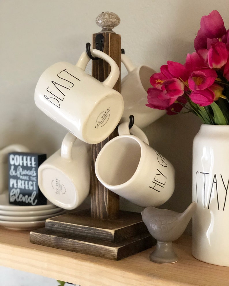 May include: A wooden mug rack with four white ceramic mugs hanging from it. The mugs have black lettering that reads "Beast", "Hey Girl", and "Stay". The rack is sitting on a wooden shelf with a stack of white plates and a small gray bird figurine.