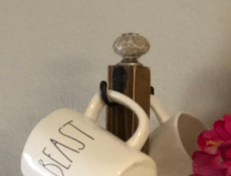 May include: A wooden mug rack with a white ceramic knob on top. The rack is holding two white mugs, one with the word "Beast" printed on it.